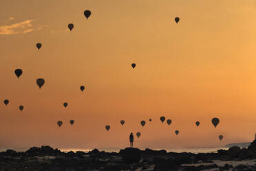 Indonesien, West Nusa Tenggara, Silhouetten von Heißluftballons fliegen über eine einsame Frau am felsigen Ufer in der stimmungsvollen Abenddämmerung - KNTF04655