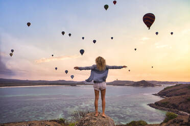 Indonesia, West Nusa Tenggara, Hot air balloons flying over lone woman standing on rocky shore with raised arms - KNTF04657