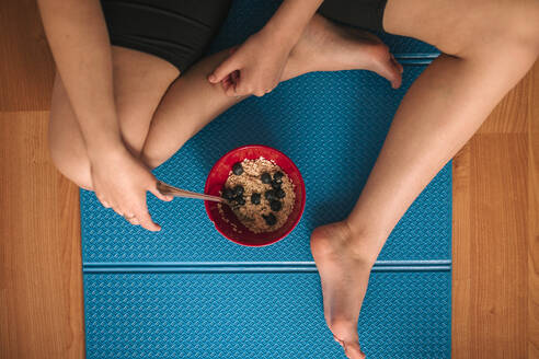 Young woman with breakfast bowl sitting on exercise mat at home - GRCF00267