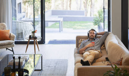 Man relaxing on living room sofa, listening to music with headphones and mp3 player - HOXF06277