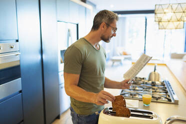 Mann liest Zeitung und bereitet Toast in der Morgenküche vor - HOXF06287