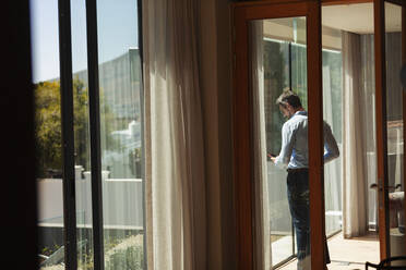 Businessman using smart phone at sunny window - HOXF06298