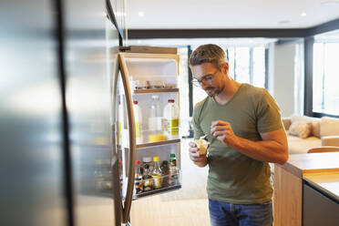 Mann isst Joghurt am offenen Kühlschrank in der Küche - HOXF06305