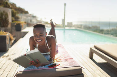 Lächelnde, unbekümmerte junge Frau, die am sonnigen Pool ein Buch liest - HOXF06337