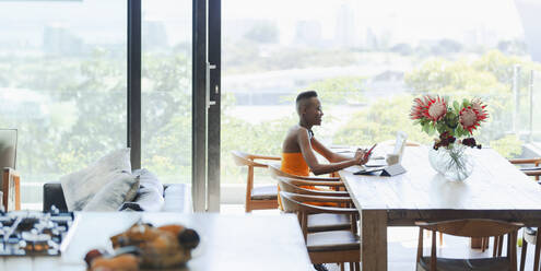 Young woman using smart phone and laptop, working from home at dining table - HOXF06347