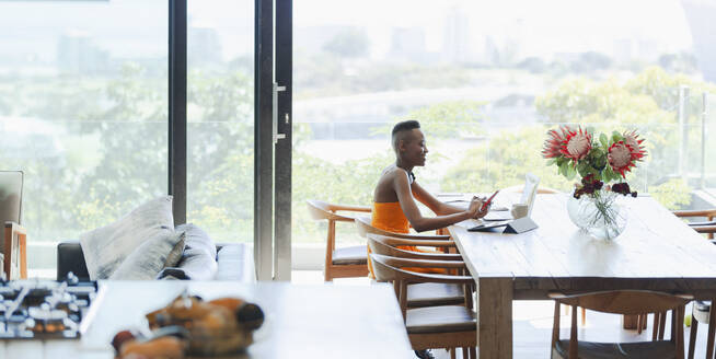 Junge Frau mit Smartphone und Laptop, die von zu Hause aus am Esstisch arbeitet - HOXF06347