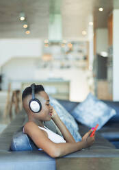 Young woman listening to music with headphones and mp3 player on sofa - HOXF06393