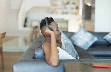 Smiling, serene young woman listening to music with headphones and mp3 player on living room sofa - HOXF06394