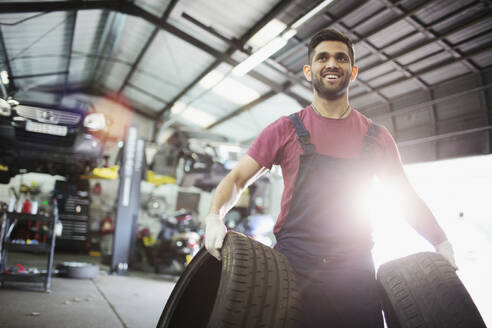 Portrait lächelnder männlicher Mechaniker, der Reifen in einer Autowerkstatt trägt - HOXF06411
