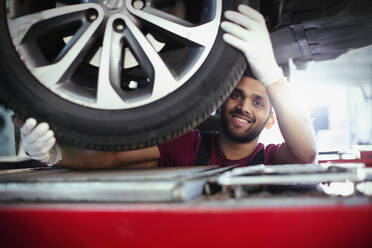Porträt eines selbstbewussten, lächelnden männlichen Mechanikers, der in einer Autowerkstatt unter einem Auto arbeitet - HOXF06433
