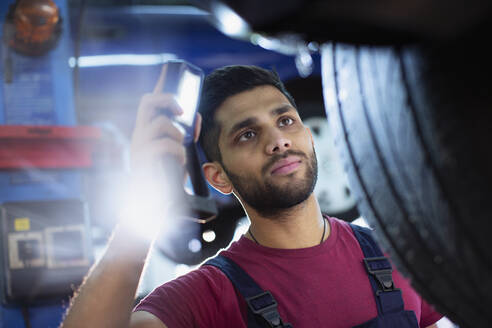 Fokussierter männlicher Mechaniker mit Taschenlampe, der einen Reifen in einer Autowerkstatt untersucht - HOXF06437