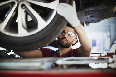 Männlicher Mechaniker, der unter dem Auto arbeitet und den Reifen in einer Autowerkstatt untersucht - HOXF06441