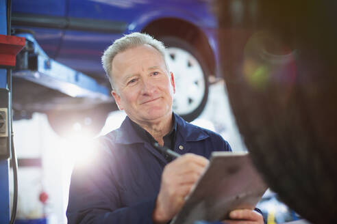 Smiling male mechanic with clipboard working in auto repair shop - HOXF06444