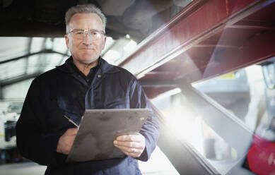 Porträt selbstbewusster männlicher Mechaniker mit Klemmbrett bei der Arbeit unter dem Auto in einer Autowerkstatt - HOXF06456