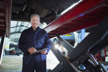 Porträt selbstbewusster, lächelnder männlicher Mechaniker mit Klemmbrett bei der Arbeit unter dem Auto in einer Autowerkstatt - HOXF06457