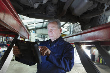 Male mechanic using diagnostic equipment under car in auto repair shop - HOXF06459