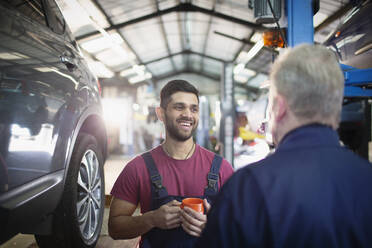 Lächelnde, glückliche männliche Mechaniker, die sich in einer Autowerkstatt unterhalten und Kaffee trinken - HOXF06461