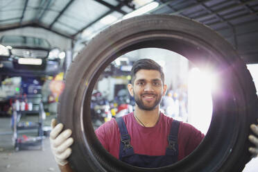 Porträt eines selbstbewussten, lächelnden männlichen Mechanikers, der einen Reifen in einer Autowerkstatt hält - HOXF06466