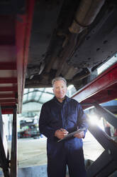 Porträt selbstbewusster männlicher Mechaniker mit Klemmbrett bei der Arbeit unter dem Auto in einer Autowerkstatt - HOXF06468