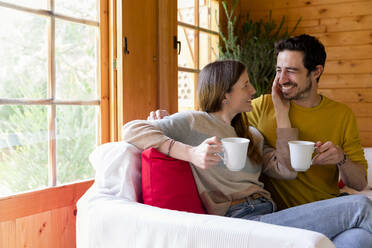 Romantic couple holding coffee mugs looking at each other while sitting on sofa in log cabin - LVVF00003