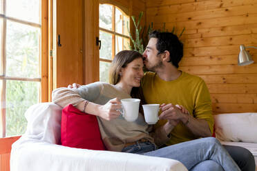 Romantischer Freund hält Kaffeetasse und küsst Frau auf die Stirn, während er in einer Blockhütte sitzt - LVVF00004