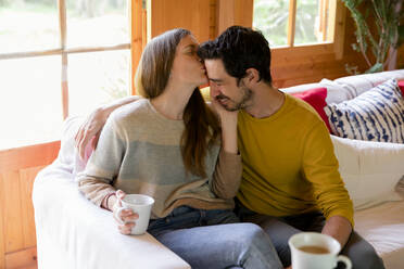 Romantic woman kissing on boyfriend's forehead while sitting over sofa in log cabin - LVVF00006