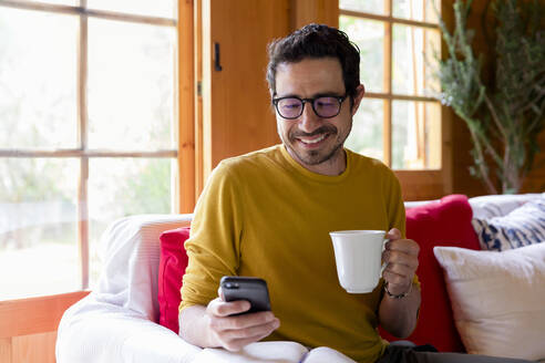 Smiling man holding coffee using smart phone while sitting on sofa in log cabin - LVVF00007