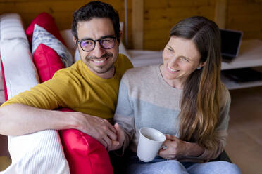 Lächelndes Paar hält sich an den Händen und entspannt sich auf dem Sofa in einer Blockhütte - LVVF00010