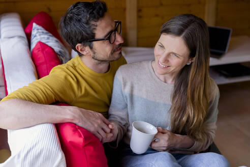 Mann betrachtet nachdenkliche Freundin mit Kaffee in der Hand, während er auf dem Sofa in einer Blockhütte sitzt - LVVF00011