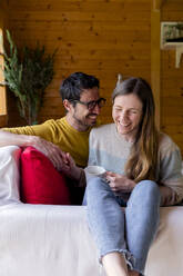 Cheerful couple holding hands while relaxing on sofa in log cabin - LVVF00012
