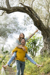 Man piggybacking cheerful girlfriend while walking against trees - LVVF00022