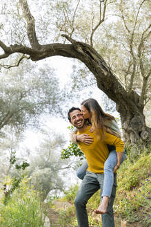 Fröhlicher Mann, der seine romantische Freundin huckepack nimmt, während er auf dem Lande gegen Bäume steht - LVVF00023
