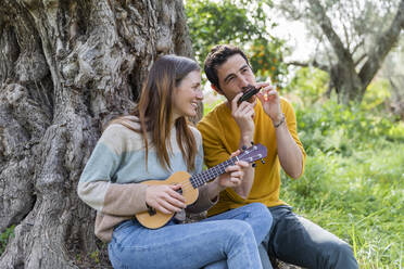 Happy couple playing musical instruments while sitting by tree trunk - LVVF00024