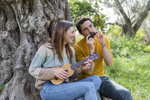 Glückliches Paar spielt Musikinstrumente, während es an einem Baumstamm sitzt - LVVF00024