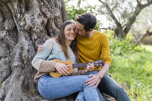 Romantischer Freund küsst glückliche Frau, die Gitarre spielt, während sie gegen einen Baumstamm sitzt - LVVF00026