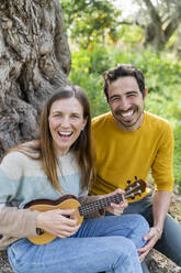 Cheerful woman playing guitar while sitting with boyfriend against tree at countryside - LVVF00027