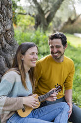 Cheerful man looking at girlfriend playing guitar while sitting in countryside - LVVF00028
