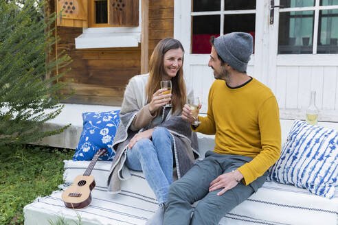 Glückliches Paar mit Weingläsern in der Hand, das sich im Sitzen an einer Blockhütte unterhält - LVVF00048