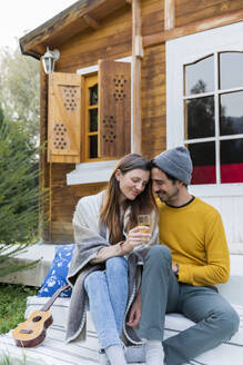 Romantischer Mann sitzt mit seiner Freundin und hält ein Weinglas vor einer Blockhütte - LVVF00052