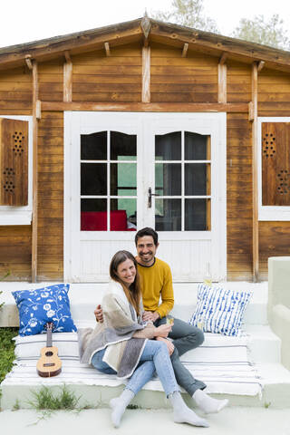 Smiling couple sitting on steps against log cabin stock photo