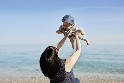 Mother picking up happy cute son against sea during sunny day - JNDF00163