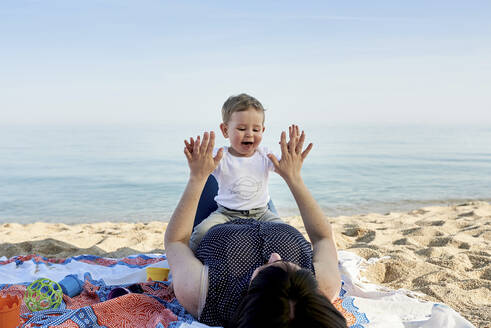 Mutter spielt mit ihrem Sohn ein Klatschspiel, während sie auf einer Decke am Strand liegt und das Meer sieht - JNDF00164