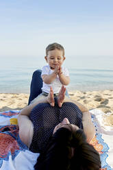 Mother playing clapping game with cute son while lying on blanket at beach against sea - JNDF00165