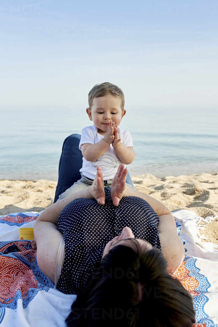 Mother playing clapping game with cute son while lying on blanket at beach  against sea stock photo