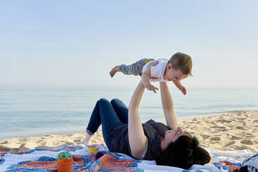 Mutter hebt glücklichen Sohn auf, während er auf einer Decke am Strand liegt - JNDF00166