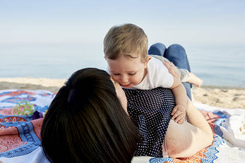 Mother kissing cute son while lying on blanket at beach against sky stock photo