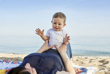 Cute smiling boy sitting on mother's stomach at beach against sky - JNDF00168