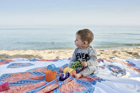 Niedlicher Junge mit Spielzeug auf einer Decke sitzend am Strand gegen das Meer, lizenzfreies Stockfoto