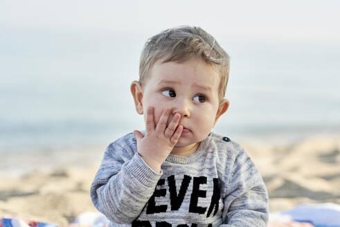 Nahaufnahme eines niedlichen Jungen mit der Hand im Gesicht am Strand sitzend gegen den Himmel - JNDF00170