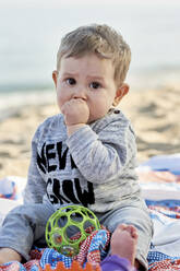 Cute boy sucking thumb while sitting on blanket at beach - JNDF00171